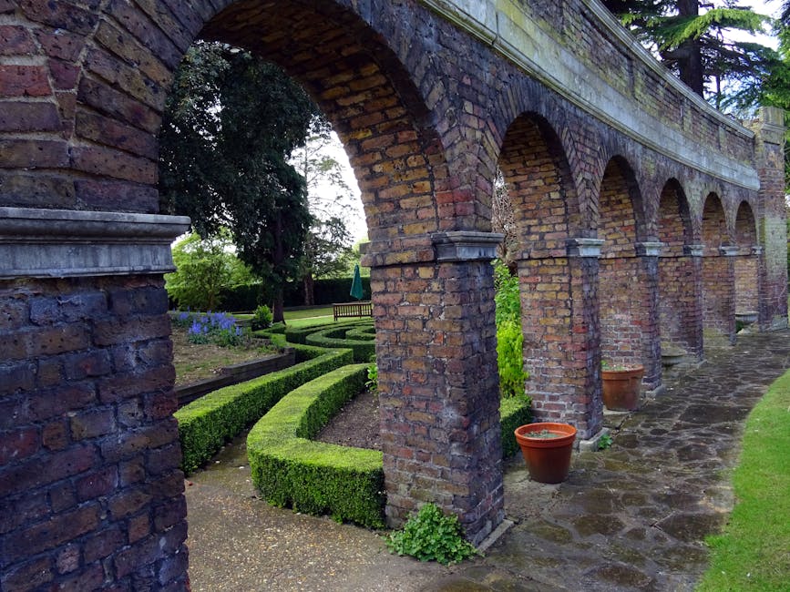 A brick garden pathway with a series of arches supported by sturdy brick columns, leading through a well-maintained landscape. To the left, neatly manicured hedges curve around flower beds filled with